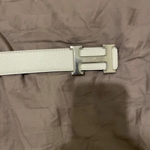  skärp med H-spänne - Säljer ett vändbart H-bälte i skinn med klassiskt H-spänne i silver. Ena sidan är vit och andra svart. Storlek 115cm.