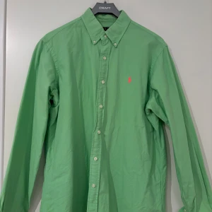 Grön skjorta från Polo Ralph Lauren - Fräsch grön skjorta från Polo Ralph Lauren med klassisk button-down krage och rosa broderad logga på bröstet. Skjortan har långa ärmar, vita knappar och är tillverkad i mjuk bomull. Perfekt för dig som gillar stilrena och färgglada plagg.