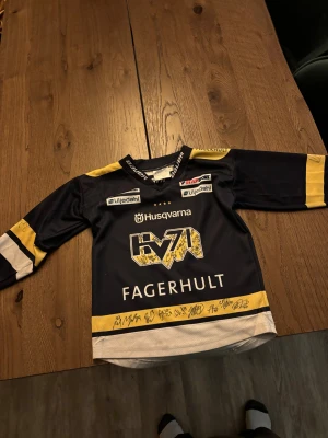 HV71 matchtröja signerad - HV71 hockeytröja med lång ärm, i mörkblått, gult och vitt. Tröjan har tryckta sponsorer och är signerad av spelare på bröstet och ärmarna. Materialet är ventilerande polyester, perfekt för hockey eller samling. Snygg och sportig design med klassiska HV71-loggan. Storlek 110-120 laget säsongen 23-24