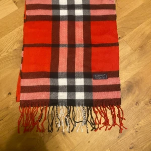 Burberrys halsduk - Snygg rutig halsduk från Burberrys i 100% lammull, tillverkad i England. Färgerna är röd, svart, vit och rosa med klassiskt rutmönster och fransar i ändarna. Perfekt accessoar för att lyfta din outfit med lite brittisk vibe.