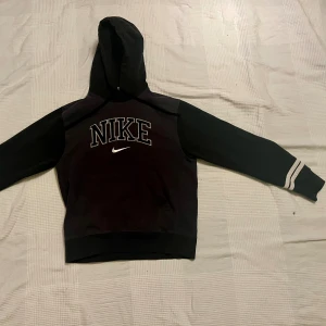 Svart hoodie från Nike med logga - Svart hoodie från Nike med stor broderad logga och swoosh på bröstet. Tröjan har huva, långa ärmar med vita ränder vid mudden och en klassisk känguruficka framtill. Perfekt för chill eller träning, riktigt snygg streetstil.