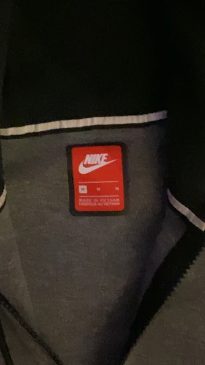 Grå hoodie från Nike med dragkedja - Snygg grå hoodie från Nike i storlek M med svart Nike-logga på bröstet. Tröjan har hel dragkedja, huva med snörning och två fickor framtill. Materialet är mjukt och känns som en blandning av bomull och polyester, perfekt för chill eller träning.