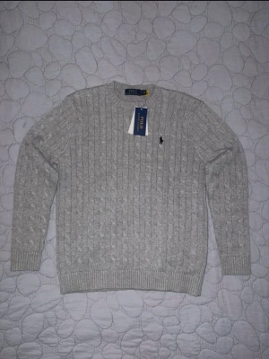Grå stickad tröja från Polo Ralph Lauren - Snygg grå stickad tröja från Polo Ralph Lauren med klassiskt kabelmönster och rund hals. Tröjan har lång ärm och den ikoniska Polo-loggan broderad på bröstet. Perfekt för dig som gillar stilrena och tidlösa plagg. Köptes i Tokyo, men var för liten för mig.