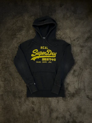 Grå hoodie från Superdry - Grå hoodie från Superdry med stor gul logga och japansk text på bröstet. Klassisk huva med snörning och känguruficka framtill. Mjuk insida och långa ärmar med muddar. Perfekt för dig som gillar streetwear och vill sticka ut med en fet logga.