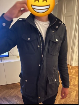 J Lindberg jacka till trendclothing - Säljer en svart parkajacka med flera fickor och snygga silverfärgade knappar. Jackan har dragkedja framtill och en hög krage som ger extra stilpoäng. Perfekt för dig som vill ha en cool och funktionell jacka med lite streetkänsla.
