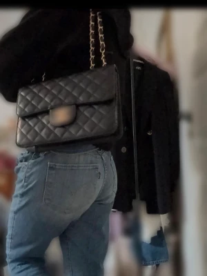 Svart axelväska med kedja - Säljer en svart axelväska med quiltat mönster och snygg kedjerem i guld och svart. Väskan har en klassisk rektangulär form och ett lock med metallspänne framtill. Perfekt för dig som gillar stilrena detaljer och vill ha en väska som sticker ut.