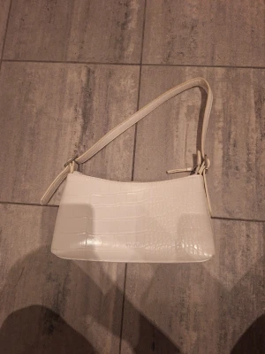 Vit axelväska från NA-KD - Säljer en stilren vit axelväska från NA-KD med croco-mönstrad yta i syntetmaterial. Väskan har justerbar rem och dragkedja upptill. Perfekt storlek för det viktigaste och har ett innerfack med dragkedja. Enkel och trendig design som lyfter din outfit.