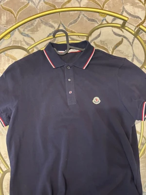 Mörkblå Moncler piké - Säljer en mörkblå pikétröja från Moncler med klassisk krage och knappar framtill. Pikén har röda och vita ränder på krage och ärmslut samt Moncler-logga broderad på bröstet. Materialet är mjuk bomull och modellen är kortärmad.