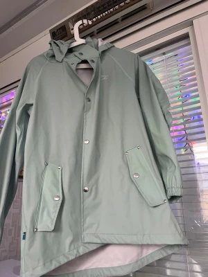 Mintgrön regnjacka med huva - Snygg mintgrön regnjacka från Didriksons med huva och tryckknappar framtill. Jackan har två stora fickor med lock och resår vid ärmsluten. Perfekt för regniga dagar och enkel att matcha med olika outfits.