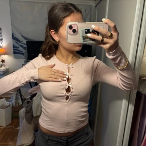 Ljusrosa ribbad topp med rosetter från Gina Tricot - Supersöt ljusrosa långärmad topp från Gina Tricot med ribbad struktur och fyra små rosetter framtill. Toppen är croppad och har en snygg detalj med öppningar längs bröstet. Perfekt att styla med jeans eller kjol för en trendig look. 