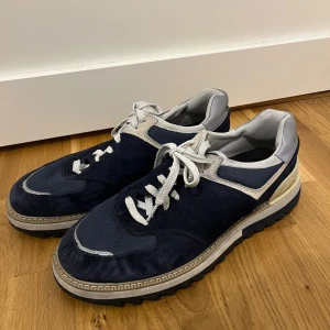 New Balance sneakers marinblå och grå - Säljer ett par New Balance sneakers i marinblått och grått med vit snörning. Skorna har en grov, räfflad platt sula och detaljer i mocka och syntet. Insidan är vadderad för extra komfort och märket syns tydligt på hälen och innersulan.