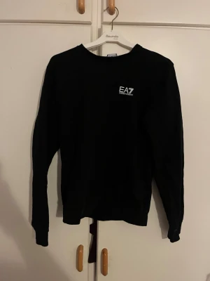 Svart sweatshirt från EA7 Emporio Armani - Säljer en svart sweatshirt från EA7 Emporio Armani med vit logga på bröstet. Tröjan har rund hals och långa ärmar, perfekt för en chill och stilren look. Materialet är mjukt och bekvämt, passar bra till jeans eller joggers.