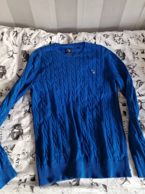Blå kabelstickad tröja från Gant - Snygg blå kabelstickad tröja från Gant med rund hals och diskret logga på bröstet. Tröjan har långa ärmar och ribbade muddar vid ärmslut och nederkant. Perfekt för dig som gillar klassisk stil med en modern touch.