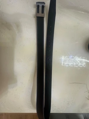 Svart skärp med metallspänne - Snyggt svart skärp med grov metallspänne i H-form. Skärpet har vit kontrastsöm längs kanterna och är gjort i ett skinnliknande material med lätt textur. Perfekt för att lyfta din outfit med en cool detalj.kan diskutera pris ganska bra.