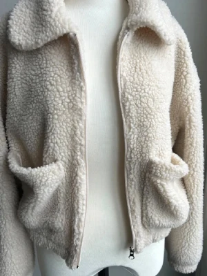 Beige teddyjacka från Pull&Bear - Mysig teddyjacka i beige från Pull&Bear med fluffig yta och klassisk krage. Jackan har två stora fickor framtill och stängs med dragkedja. Perfekt för dig som vill ha en chill och trendig look under kyliga dagar.