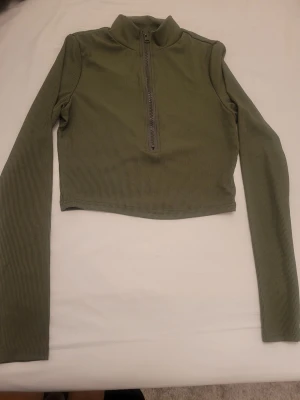 Olivgrön croppad polotopp från SHEIN - Säljer en olivgrön croppad polotopp från SHEIN i ribbad polyester. Toppen har lång ärm, hög krage och en snygg half zip-dragkedja framtill. Perfekt till jeans eller kjol för en trendig look.