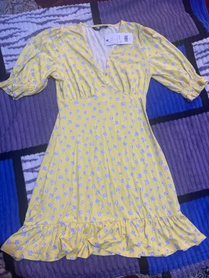 Gul blommig klänning French Connection - Supersöt gul klänning från French Connection med lila blommönster, v-ringad hals och volangkant nedtill. Klänningen har korta puffärmar med resår och är tillverkad i mjuk viskos med lite stretch. Perfekt för dig som gillar färg och mönster!