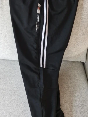 Svarta träningsbyxor med vita ränder - Svarta träningsbyxor från REC med vita ränder längs sidorna och tryckt text på låret. Byxorna har muddar vid bensluten och är tillverkade i 95% polyester och 5% elastan, vilket gör dem mjuka och stretchiga. Perfekta för sport eller chill.