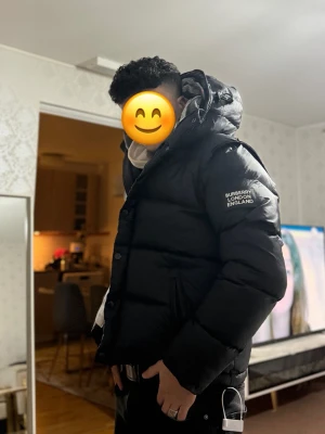 Svart pufferjacka från Burberry - Säljer en svart pufferjacka från Burberry med tydlig logga på ärmen och klassiskt rutigt foder i huvan. Jackan har dragkedja och knappar framtill samt två fickor. Perfekt för kalla dagar och riktigt snygg streetstil.