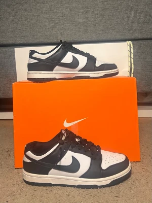 Nike Dunk Low svart och vit - Nike Dunk Low sneakers i svart och vit med klassisk låg siluett. Skorna har svart snörning, rund tå och platt sula. Ovandelen är i skinn med perforerad tåbox för extra ventilation. Swoosh-loggan syns tydligt på sidorna och de har en stilren kontrast mellan svart och vitt.