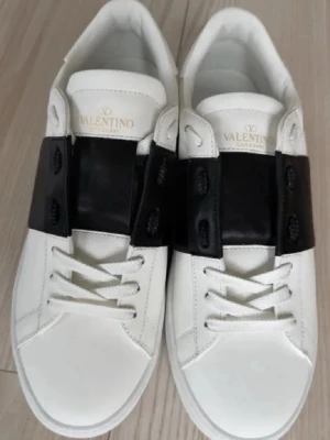 Valentino Garavani Open sneakers vit/svart - Säljer ett par snygga Valentino Garavani Open sneakers i vitt skinn med bred svart läderdetalj över snörningen. Skorna har rund tå, platt sula och coola nitar på hälen. Perfekt för dig som gillar exklusiva märken och stilren design.