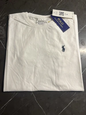 Vit t-shirt från Polo Ralph Lauren - Vit t-shirt från Polo Ralph Lauren med klassisk rund hals och korta ärmar. T-shirten har den ikoniska Polo-loggan broderad i mörkblått på bröstet. Tillverkad i mjuk bomull för en skön och avslappnad känsla. Perfekt för dig som gillar stilrena och tidlösa plagg.
