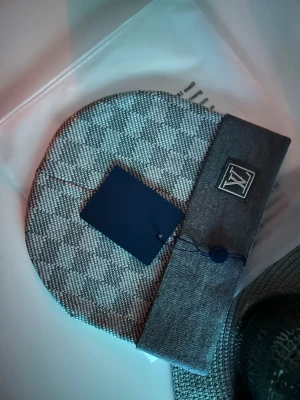 Grå rutig Louis Vuitton väst - Säljer en riktigt snygg väst från Louis Vuitton i grått med rutigt mönster. Västen har en bred enfärgad kant nertill och en diskret LV-logga på sidan. Materialet känns mjukt och exklusivt, perfekt för dig som vill sticka ut med stil.