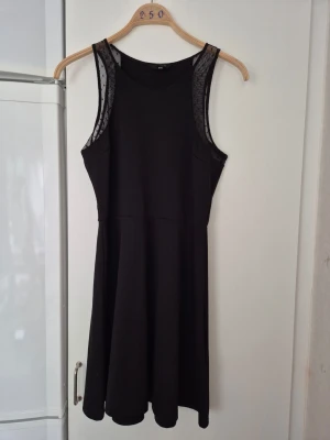 Klänning - Säljer en svart klänning från H&M i storlek M. Klänningen har ärmlös design med meshdetaljer upptill och en lätt utställd kjol. Perfekt för dig som gillar enkel men snygg stil.