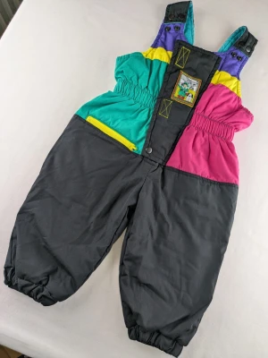 Färgglada retro täckbyxor med brodyr - Unika täckbyxor i retrostil med färgblock i svart, lila, gul, turkos och rosa. Byxorna har resår i midjan och benslut, justerbara axelband med tryckknappar, gul dragkedja och en broderad patch framtill. Perfekta för vinterns alla äventyr. Some wear on the back. Please see last photo.