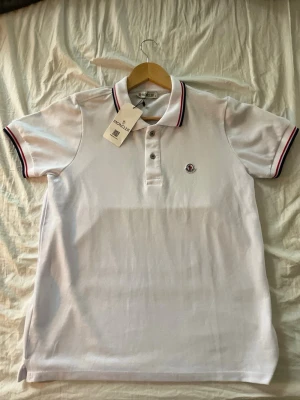 Vit Moncler piké med rödblå detaljer - Säljer en fräsch vit pikétröja från Moncler i storlek S. Tröjan har korta ärmar, klassisk krage med röd och blå rand, samt Moncler-logga broderad på bröstet. Materialet är bomull och knäppningen framtill har tre knappar. Perfekt för dig som gillar stilrena märkesplagg. Pris går att diskuteras!