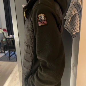 Parajumper cardigan  - Säljer en mörkgrön cardigan från Parajumpers med huva och coolt patch på ärmen. Jackan har quiltad framsida och rygg, meshade ärmar och sidopaneler, samt dragkedja framtill med gul detalj vid halsen. Storlek L men passar även som M. Vid snabb affär kan priset förhandlas om 
