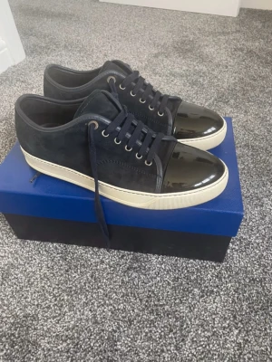 Mörkblå sneakers med lackad tå - Snygga mörkblå sneakers med snörning och vit sula. Skorna har en unik lackad tå i svart som ger en cool kontrast mot den matta mockan på resten av skon. Perfekt för dig som gillar stilrena detaljer och vill sticka ut lite extra.