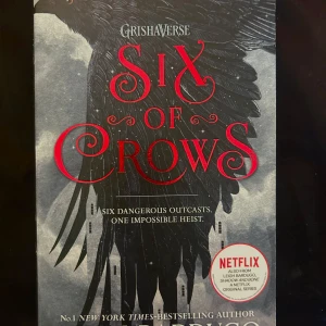 Six of Crows - Säljer boken 'Six of Crows' från Grishaverse-serien. Omslaget är svart och grått med röda detaljer och motiv av en kråka. Boken är på engelska och är första delen i en duologi. Perfekt för dig som gillar fantasy, äventyr och spänning.