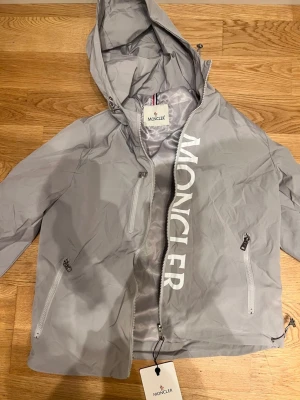Moncler windbreaker - Säljer en grå vindjacka från Moncler med stor logga framtill och klassisk patch på ärmen. Jackan har huva med dragsko, flera dragkedjeförsedda fickor och hel dragkedja framtill. Perfekt för dig som vill ha en clean och sportig look.