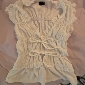 Vit omlottopp med knyt från Gina Tricot - Supersöt vit omlottopp från Gina Tricot i storlek XXS. Toppen har volangdetaljer, knyt framtill och är ärmlös. Inga defekter ❤️