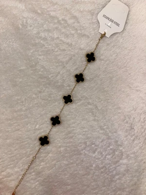 Svart och guldigt armband i stål - Snyggt armband i rostfritt stål med fem svarta fyrklöverformade hängen och en i silver, alla infattade i guldfärgad ram. Kedjan är tunn och justerbar för perfekt passform. Perfekt accessoar för att lyfta din outfit.