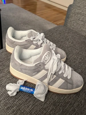 Adidas Campus grå sneakers strl 42 - Säljer ett par Adidas Campus sneakers i grå mocka med vita detaljer och klassiska tre ränder på sidan. Skorna har rund tå, platt sula och snörning. Adidas-logga på plösen och extra vita skosnören medföljer. Perfekt för dig som gillar stilrena och tidlösa sneakers.