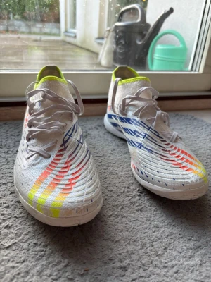 Adidas vita fotbollsskor med färgglada detaljer - Säljer ett par Adidas fotbollsskor i vitt med coola mönster i blått, rött, orange och gult. Skorna har snörning, platt sula och rund tå. Insidan är neonfärgad och ovandelen är i syntetmaterial. Perfekt för dig som vill sticka ut på planen!