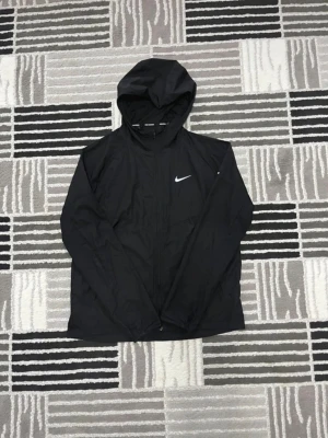 Svart Nike vindjacka - Svart vindjacka från Nike med huva och hel dragkedja framtill. Jackan har en stilren design med Nike-logga på bröstet och texten 'NIKE RUNNING' på insidan av kragen. Tillverkad i lätt polyester, perfekt för löpning eller chill dagar.