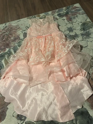 Ljusrosa långklänning med volang - Säljer en ljusrosa långklänning med glittriga detaljer och volanger. Klänningen har ett lager av transparent tyg med blommigt mönster och en söt rosett i midjan. Materialet känns som satin och tyll, perfekt för speciella tillfällen.Den är för liten för mig jag har växt och pris kan diskuteras☺️