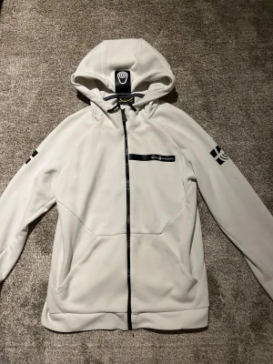 Vit hoodie från Sail Racing med luva. - Snygg vit hoodie från All Racing med svart dragkedja och coola svarta tryck på ärmarna. Tröjan har luva med snörning och praktiska fickor framtill. 