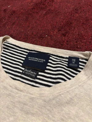 Beige tröja Scotch & Soda M - Snygg beige tröja från Scotch & Soda i storlek M. Tillverkad i en mjuk mix av kashmir och bomull. Tröjan har rund hals och långa ärmar, med diskreta ribbade muddar. Knappt använd. Priset är ej hugget i sten, hör av er vid funderingar!