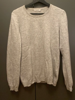 Grå kashmirtröja från J.Lindeberg - Mjuk och stilren grå tröja från J.Lindeberg i 100% kashmir. Rund halsringning och långärmad modell med ribbade muddar vid ärmslut och nederkant. Perfekt för dig som gillar enkel och klassisk design med lyxigt material. Har ett litet hål i högerarmbåge. Är i storlek L men sitter som Xs
