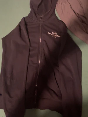 Svart Peak Performance väst med huva - Svart väst från Peak Performance med huva och hel dragkedja framtill. Diskret logga broderad på bröstet i vitt. Västen är tillverkad i mjukt material och har en sportig look, perfekt för dig som gillar stilren design och funktion.