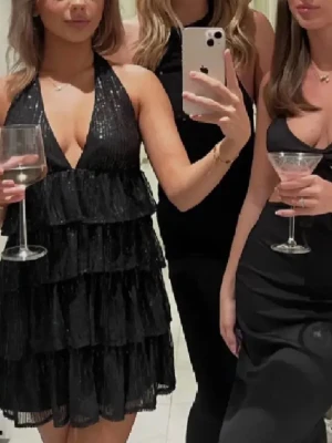 Svart glittrig volangklänning - Supersnygg svart kortklänning med glittriga paljetter och flera lager volanger. Klänningen har djup v-ringning, öppen rygg och knyt i nacken. Perfekt till nyår! 