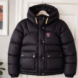 Svart pufferjacka från Fjällräven - Säljer en svart pufferjacka från Fjällräven med huva och gula snören. Jackan har två stora fickor med knappar framtill, dragkedja och tryckknappar. Klassisk Fjällräven-logga på bröstet och quiltad design som håller värmen under kalla dagar.