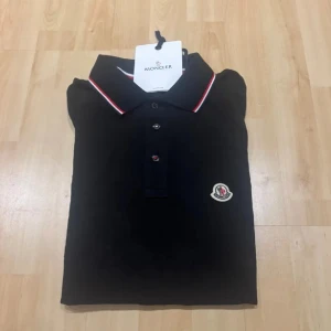 Svart pikétröja från Moncler - Svart pikétröja från Moncler med klassisk krage och knäppning framtill. Tröjan har Moncler-logga på bröstet och röda, vita och blå detaljer på ärmsluten och kragen. Perfekt för en stilren och sportig look.