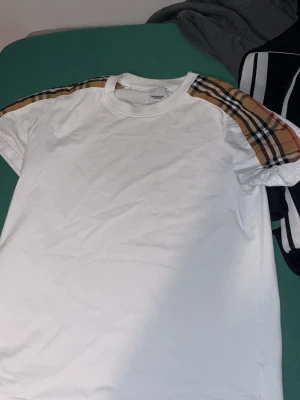 Vit Burberry t-shirt med rutiga axlar - Säljer en fräsch vit t-shirt från Burberry med klassiska rutiga paneler i beige, svart och rött på axlarna. T-shirten har rund hals och korta ärmar, perfekt för dig som gillar stilrena designer med en touch av lyx. Materialet är mjuk bomull.