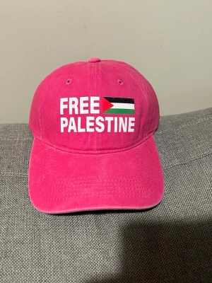 Rosa keps med texten Free Palestina.  - Rosa keps med böjd skärm och justerbart spänne bak. Framsidan har ett tryck med texten 'FREE PALESTINE' och en flagga i rött, grönt, vitt och svart. Kepsen är tillverkad i bomull och har en klassisk, avslappnad look som passar till streetwear.FREE PALESTINE KEPS - GÅRDA WASHED  DEN ÄR HELT ROSA MEN SISTA TVÅ BILDRER BARA VISAR HUR DEN SER UT. Detaljinformation:  Tillverkad av 100 procent bomull One Size Justerbar vid kepsens baksida Gjord av: 100% bomull  