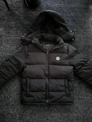 Trapstar vinterjacka  - Trapstar pufferjacka i svart med stor logga på ryggen och rund emblem på bröstet. Jackan har huva, dragkedja framtill och två sidofickor. Materialet är polyester och jackan har en loose passform som ger en chill vibe. Perfekt för kalla dagar och streetwear-stil. Storlek L men passar även XL, skick 8/10 med en liten skada vid ena fickan men inget som man tänker på. Pris går att diskutera✅